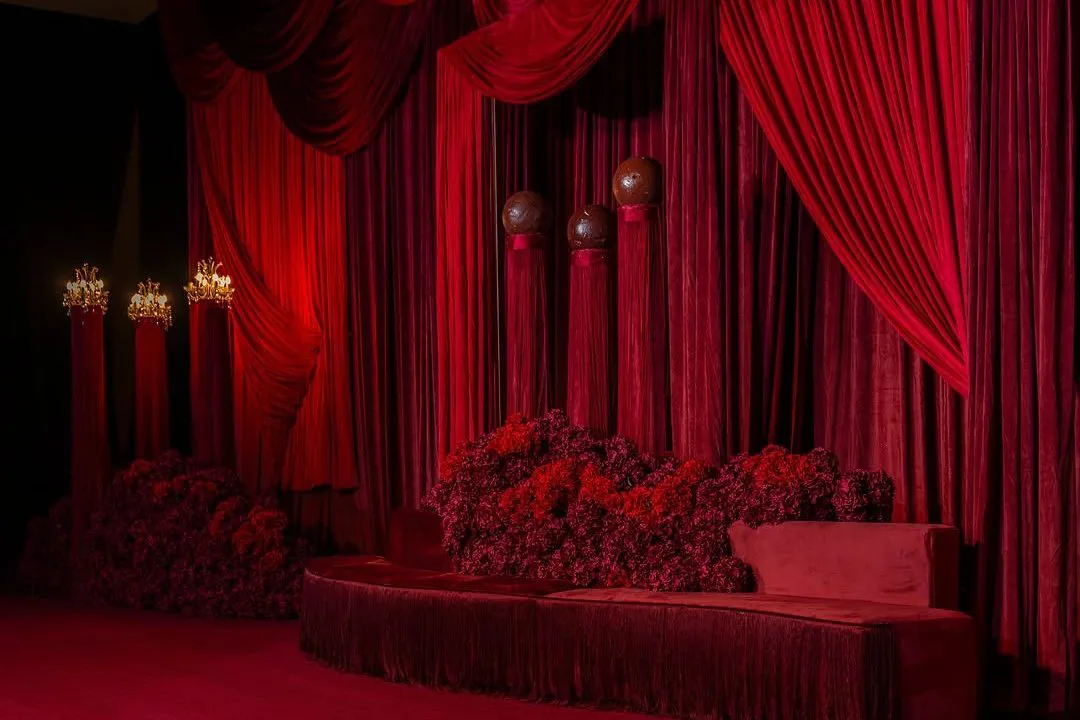 Wedding stage with a fringed velvet sofa, draped red curtains and a wall of red flowers
